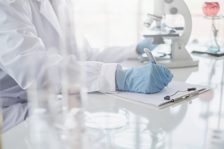 A scientist hands writing on a clipboard in laboratory with test tube microscope and solutions.